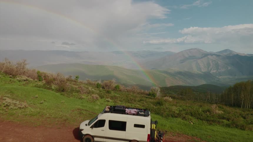 An aerial view slowly pushes past a camper van parked on a remote dirt road, revealing a vast mountain landscape with a vibrant rainbow in the sky