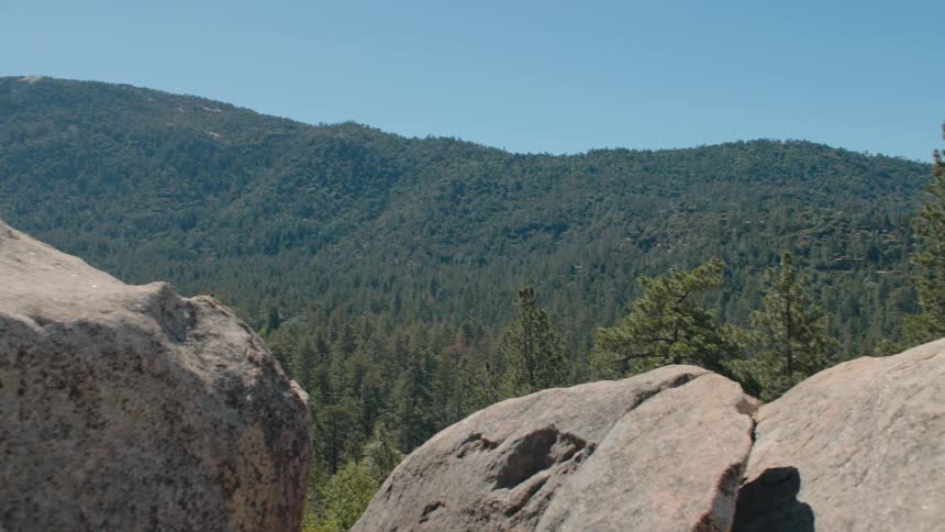 Peeking through large rocks at a vast, dense forest covering mountains under a clear blue sky