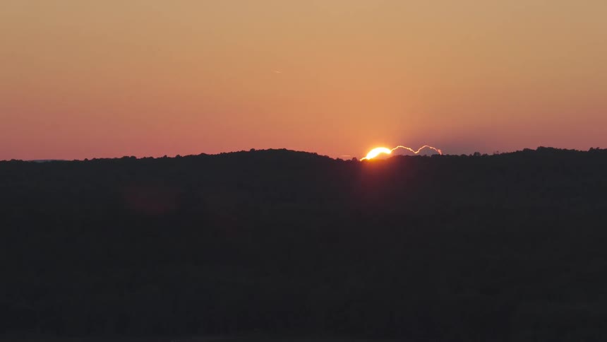 A time-lapse captures the sun peeking over a silhouetted treeline as its rays illuminate the edge of a small cloud