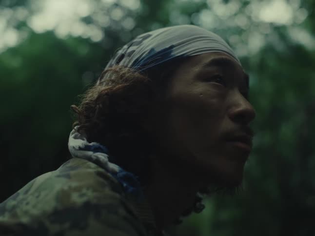 A pensive man with a bandana and camouflage shirt looks up thoughtfully in a dark, lush forest