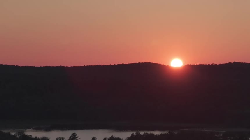 A time-lapse shows the sun rising over a silhouetted, tree-covered hill with a lake and mist in the valley below