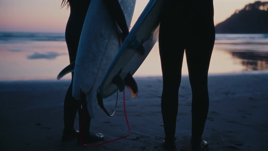Two silhouetted surfers in wetsuits stand with their boards on a sandy beach at twilight, watching the ocean