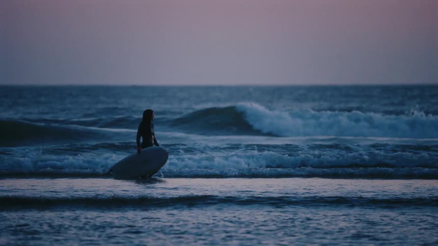 A silhouetted surfer with a board wades into the ocean as waves break in slow motion during twilight