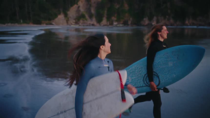 Two happy female surfers in wetsuits run along the wet sand with their boards, excited to catch some waves at dusk