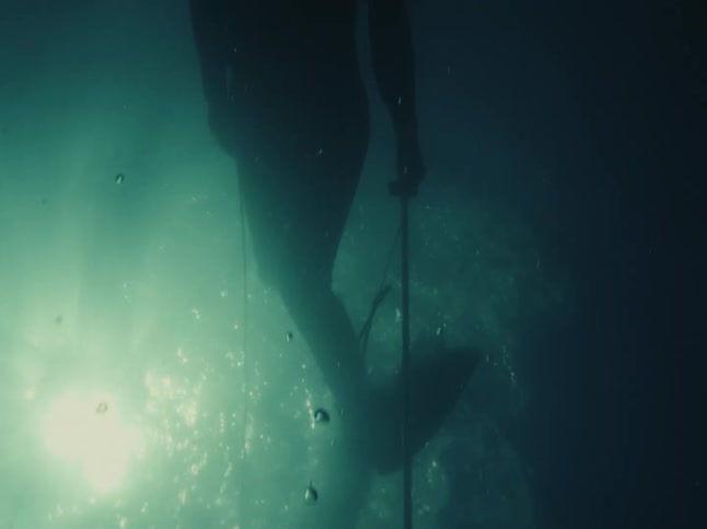 A lone free-diver holding a speargun descends into the deep, dark ocean as bubbles float to the sunlit surface