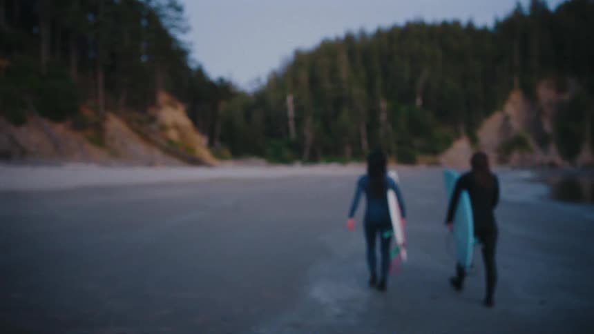 A smiling couple in wetsuits carry their surfboards and walk along a remote, sandy beach at dusk
