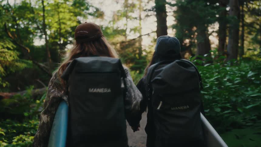 Two female surfers with backpacks carry their boards while hiking down a lush forest trail at golden hour