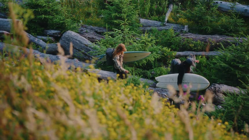 Two adventurous surfers hike through a beautiful, rugged forest with their boards and backpacks