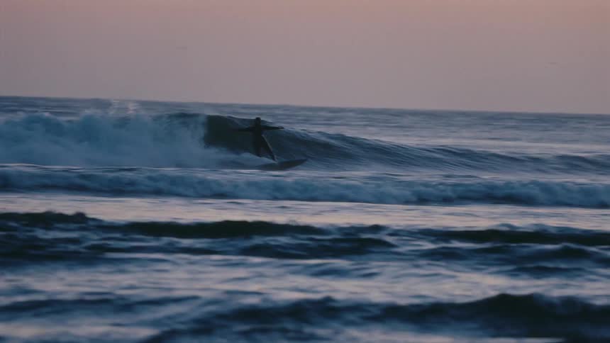A surfer skillfully rides an ocean wave in slow motion, silhouetted against the soft light of dusk