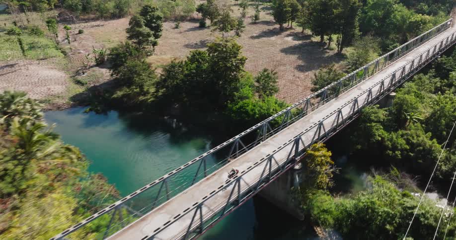 High-angle drone shot follows a person riding a motorcycle across a scenic bridge over a turquoise river in the countryside