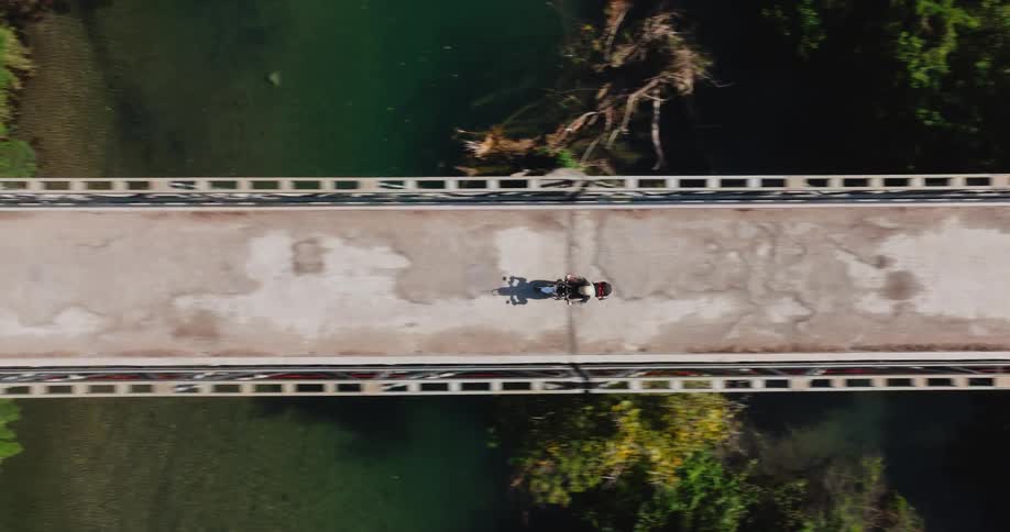 A drone follows a motorcycle from a top-down perspective as it crosses a concrete bridge over a river in the jungle