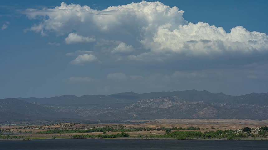 A tranquil landscape shows a massive white cumulus cloud floating over an arid mountain range and a calm lake