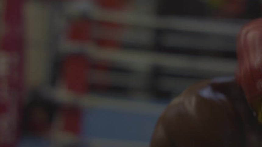An intense, shirtless boxer with red gloves practices on a speed bag in slow motion inside a dimly lit gym