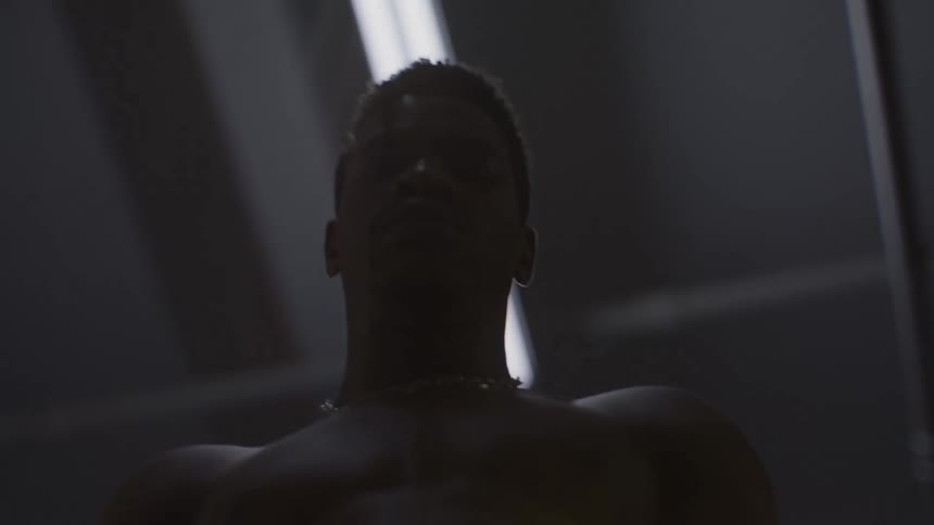 A powerful, shirtless man slowly rises into the frame from a low angle under a stark overhead light