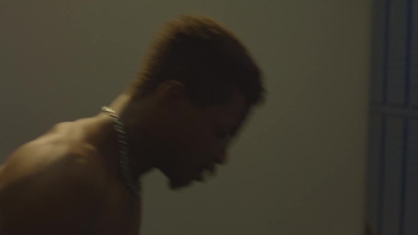 A shirtless, athletic Black man with a muscular back mentally prepares himself in a dimly lit locker room