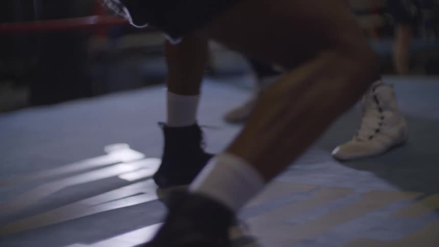 The feet of two boxers in black and white boots dance around the canvas of a boxing ring in slow motion