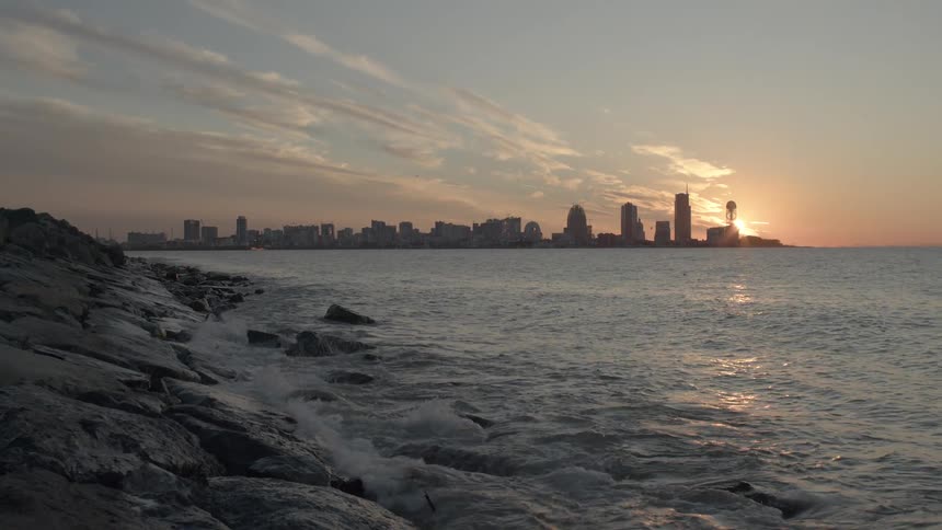 A slow pan reveals a modern city skyline across the sea from a rocky shore during a beautiful, calm sunset