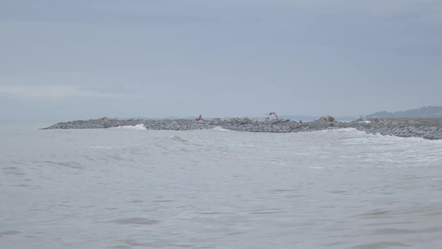 A wide pan across a turbulent gray sea with distant cargo ships reveals waves crashing against a rocky breakwater under construction