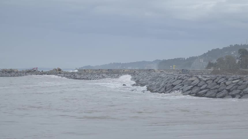 Powerful storm waves crash against a stone breakwater in slow motion on a gray, overcast day
