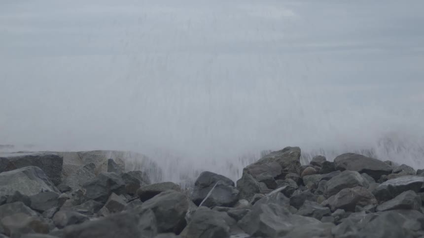 A powerful ocean wave crashes against a rocky shore in dramatic slow motion on a gray, overcast day