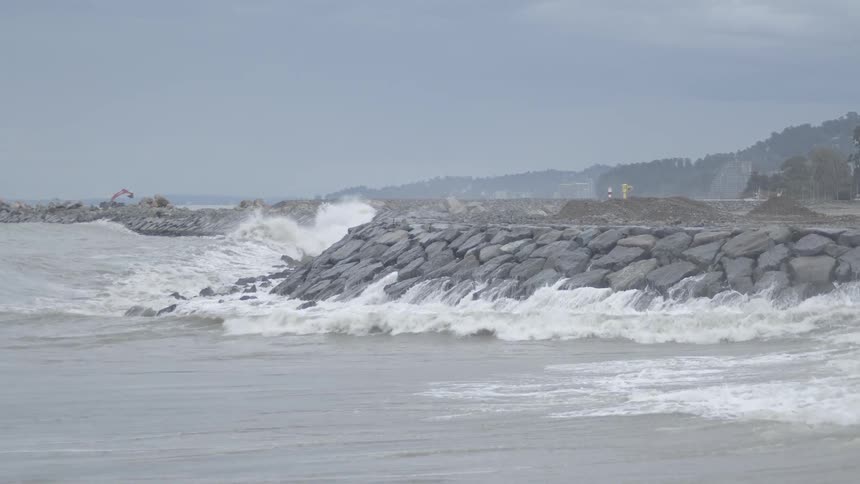 A powerful storm surge reveals massive ocean waves crashing in slow motion against a rocky breakwater on an overcast day