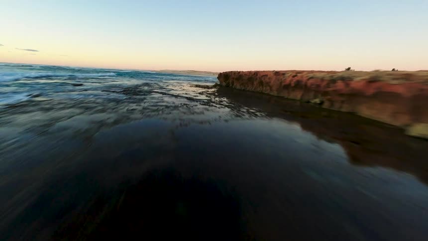 A fast-paced, low-angle drone shot skims over the ocean surface along a rugged, rocky coastline at sunset