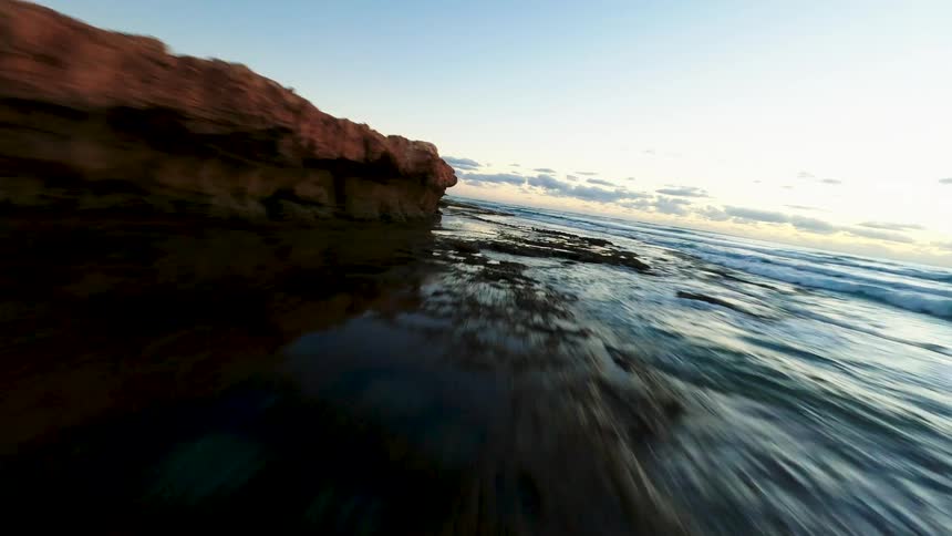 An FPV drone soars quickly along a rugged coastline, skimming just above the ocean waves and wet sand at sunrise