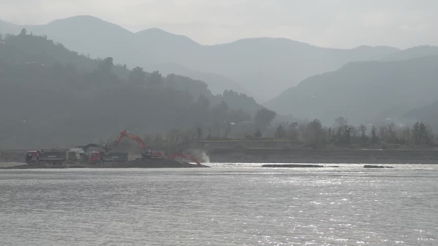 Industrial excavators and dump trucks work on a riverbank with a backdrop of distant, hazy mountains