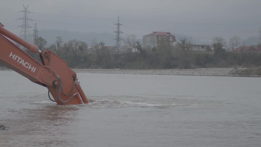 An excavator bucket scoops rocks and gravel from the bottom of a murky river in slow motion on an overcast day