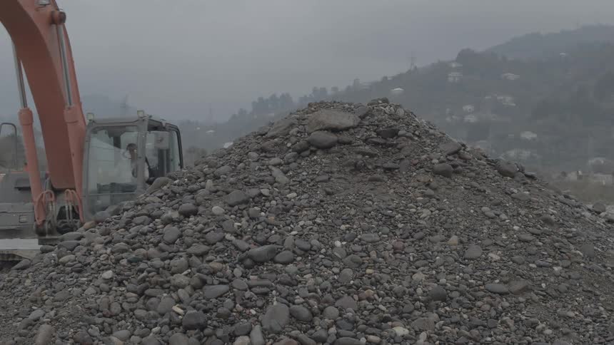 A large excavator drops a bucket full of wet gravel and sand onto a large pile at a construction site on a foggy day