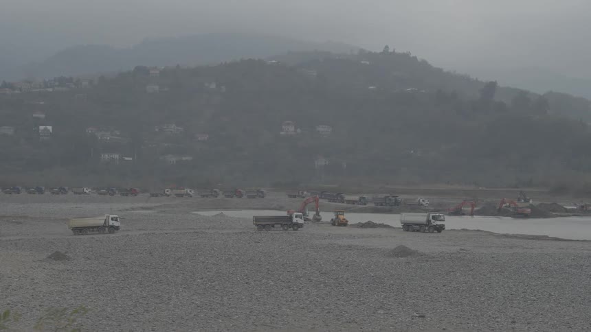 A long convoy of dump trucks waits in a vast, foggy riverbed quarry as excavators mine for sand and gravel