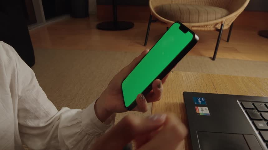 A person holds and taps on a smartphone with a green screen while sitting indoors with a laptop nearby