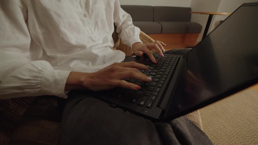 A close-up view of a person in a white blouse typing on a laptop while sitting in a wicker chair