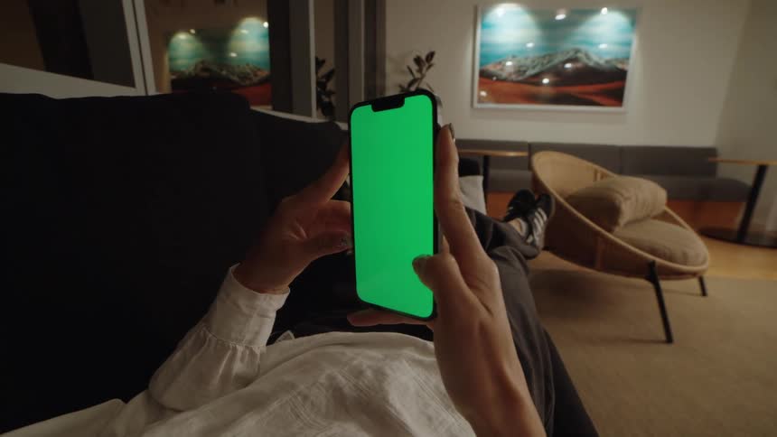 POV shot of a person lying on a couch at night and scrolling through a smartphone with a green screen