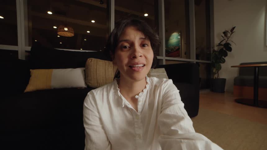 An expressive woman makes a range of facial expressions while talking to the camera during a video call from her living room at night