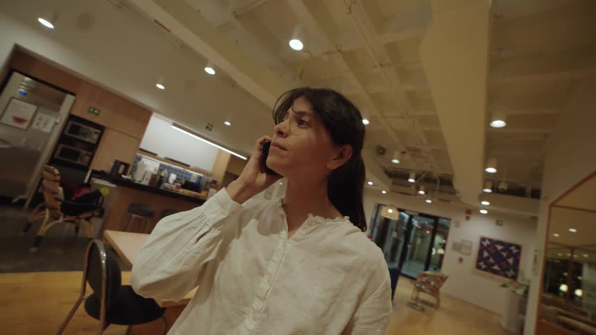 Low-angle shot of a smiling woman in a white shirt talking on her smartphone in a modern office breakroom