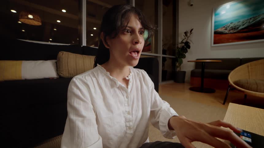 A woman sitting on the floor in a modern living room at night has a surprising conversation during a video call on her laptop