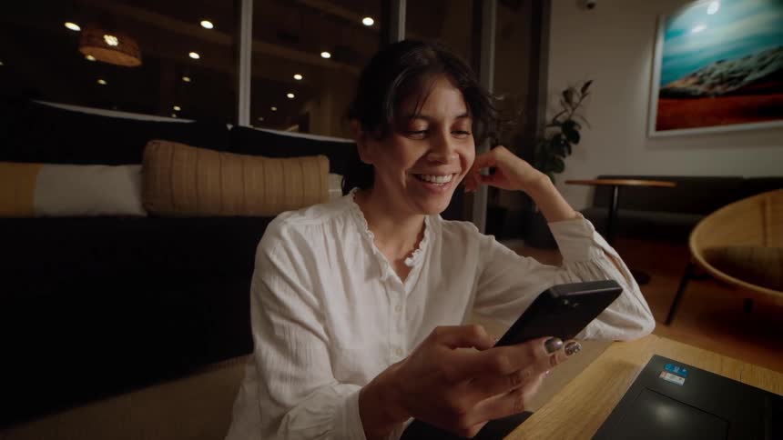 A smiling woman in a white blouse enjoys content on her smartphone while sitting in a modern, warmly lit lounge at night