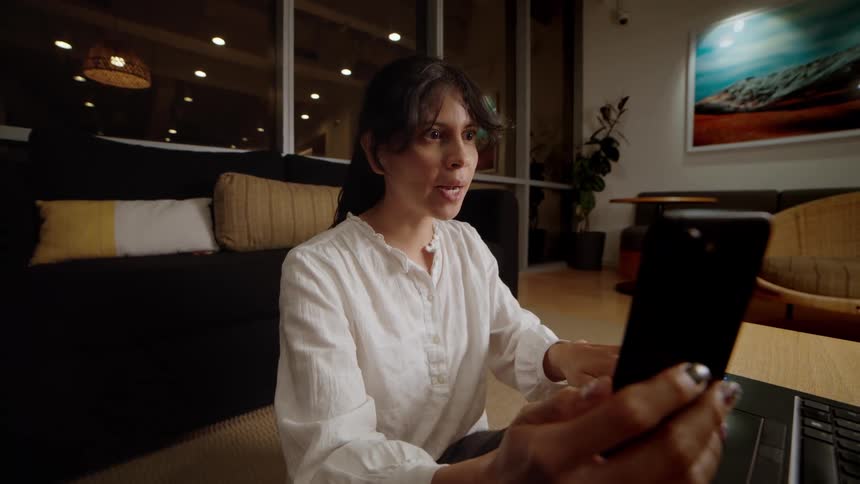 Woman multitasking on a video call from her phone while working on her laptop from a cozy, modern lounge at night