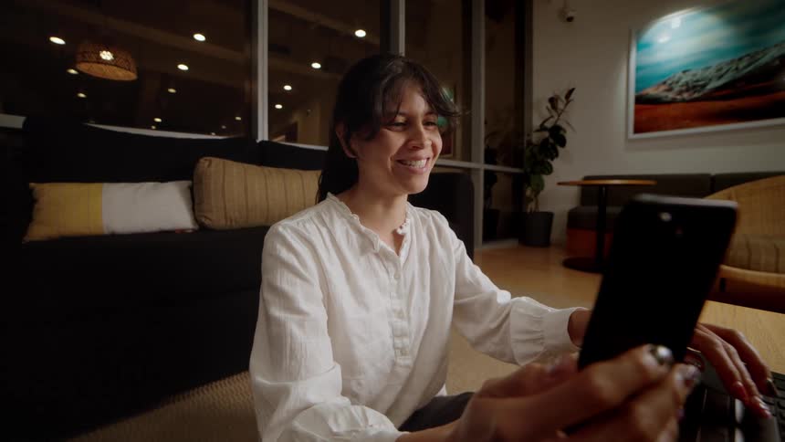 Happy woman video calling on her smartphone and working on her laptop at night in a stylish lounge