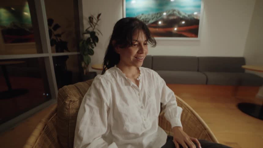 A smiling woman in a white blouse works on her laptop at night, sitting comfortably in a modern wicker chair