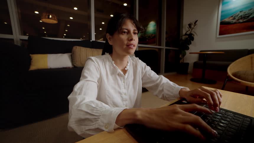 A woman sits at a low coffee table in a modern living room at night, smiling and then concentrating as she types on her laptop