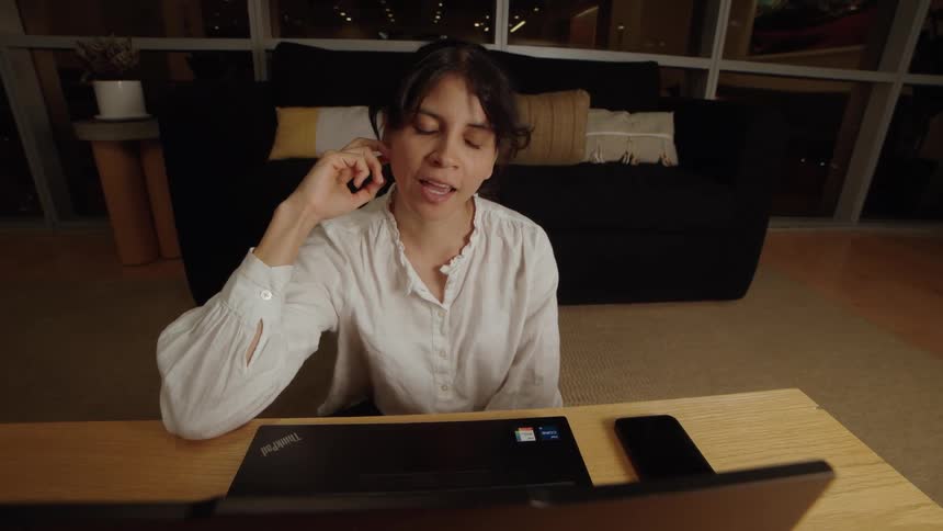 A pleasant woman smiles and chats during a video call on her laptop from her living room at night