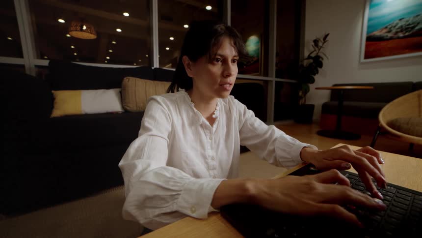 A focused woman types on a laptop at a coffee table in a modern living room at night
