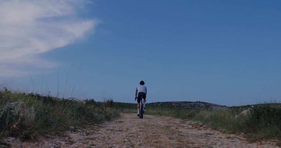 Following a lone cyclist from behind as they pedal uphill on a rural, unpaved road