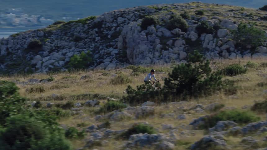 A cyclist rides his gravel bike along a rugged, rocky path on a hillside overlooking the sparkling sea