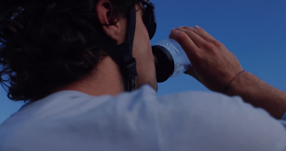 A cyclist wearing a helmet and sunglasses takes a break to drink water from his bottle against a blue sky in slow motion