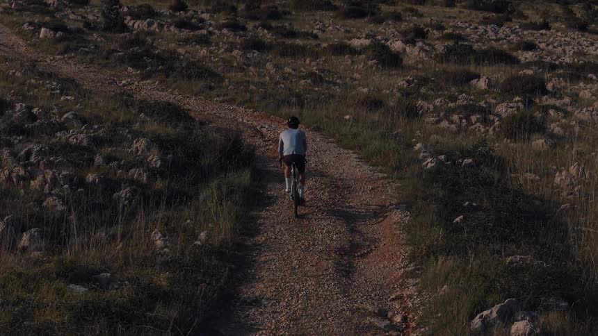 Aerial view of a lone cyclist pedaling up a rugged gravel path through a vast, rocky landscape at golden hour