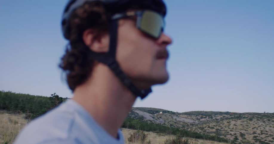 Cinematic slow-motion close-up reveals a focused male cyclist in a helmet and sunglasses looking out over the hilly landscape