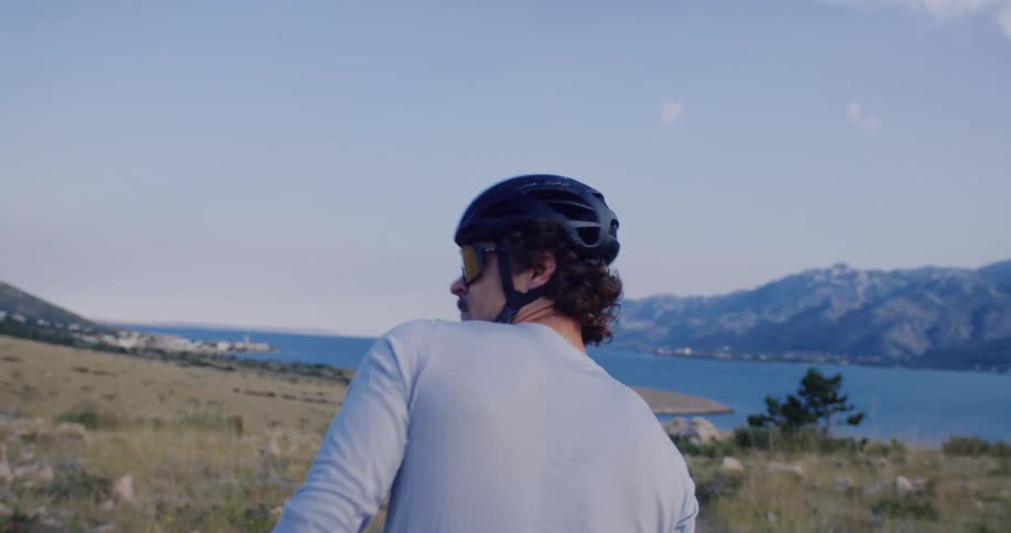 A mustached cyclist looks back over his shoulder before riding his bike down a scenic gravel path overlooking a tranquil blue bay and distant mountains